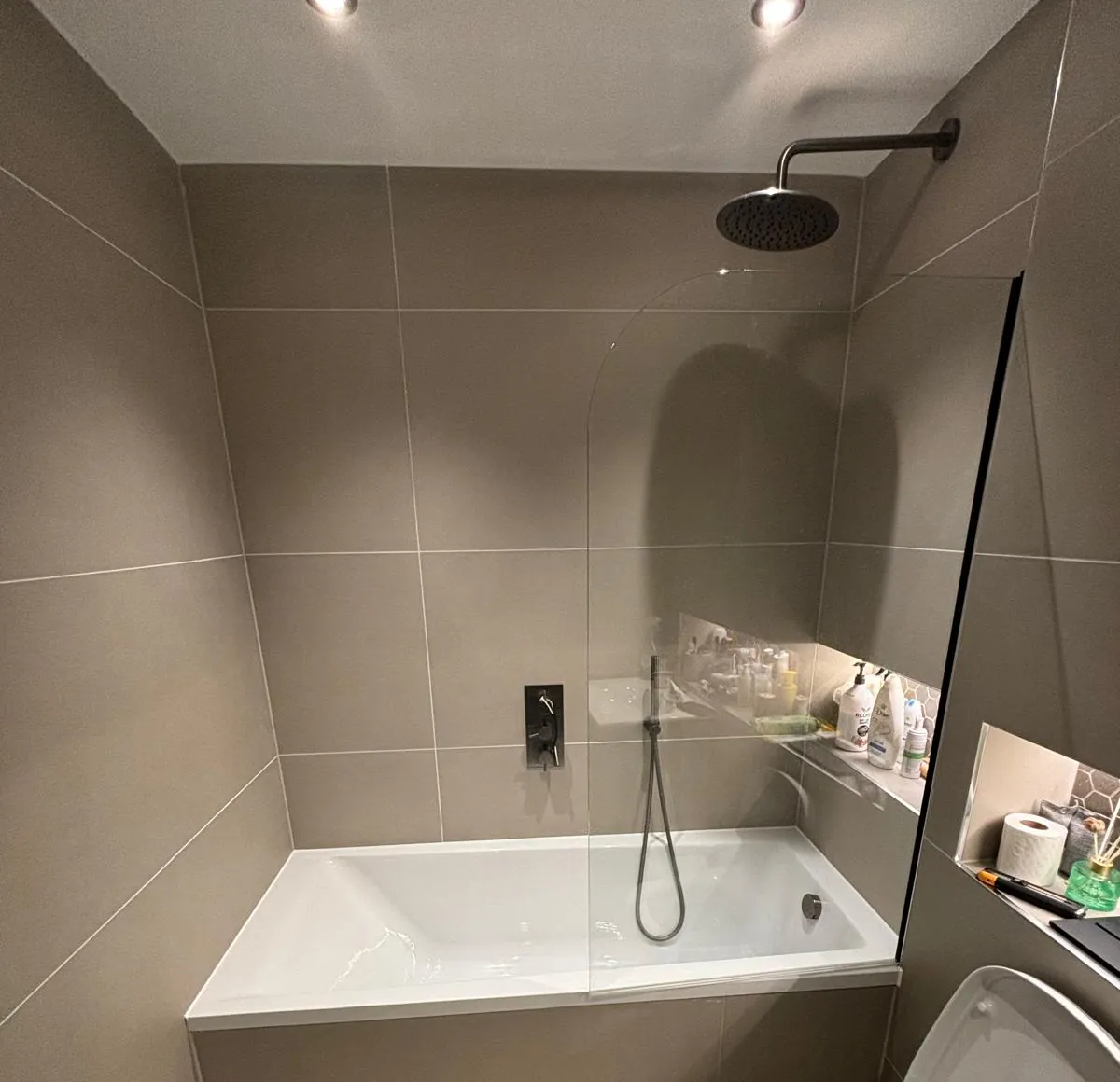 A finished grey-tiled bathroom with a bath, glass screen and black rain shower