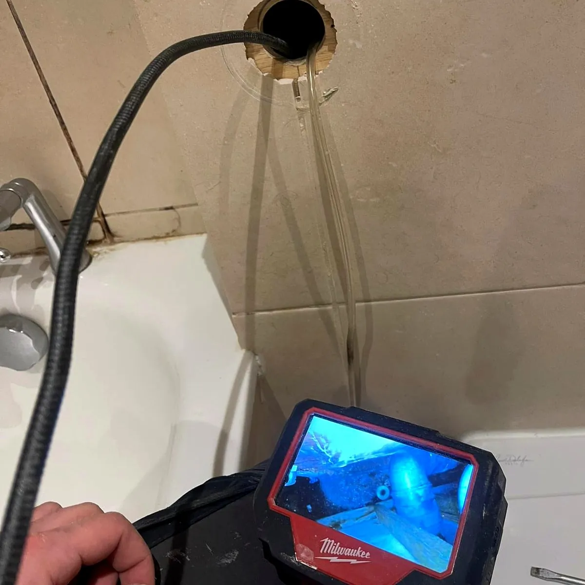 An inspection camera fed through a small hole in the tiled wall beside a bath, with its screen showing the space inside